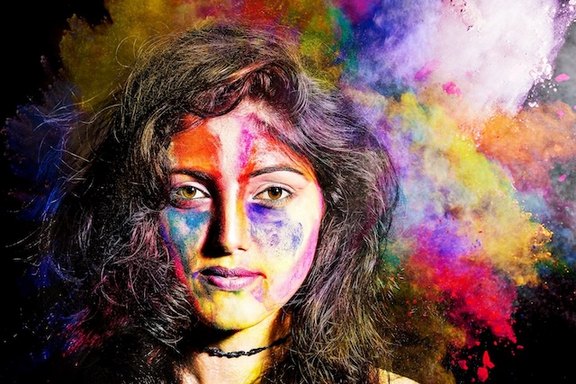 Eine junge Frau ist mit bunten Farben angemalt und steht vor einem dunklen Hintergrund mit vielen bunten Farben.