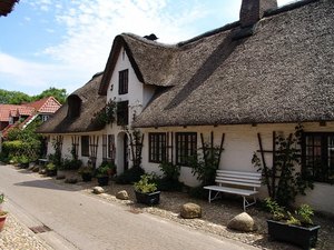 Zusehen ist ein langgestricktes älteres weißes niedriges Gebäude mit Reetdach an einer Landstraße