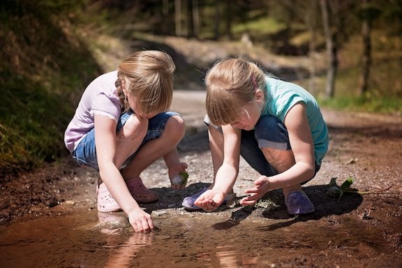 Zwei Mädchen hocken an einem Wasser und untersuchen, was sie dort am Wasserrand finden. Im Hintergrund ist ein Waldweg zu sehen.
