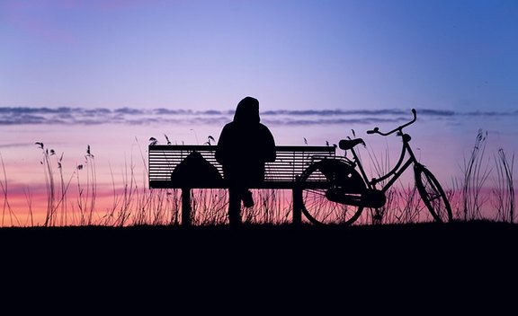 Es sitzt jemand alleine auf einer Bank am Meer und schaut in den rotgefärbten Abendhimmel, ein Fahrrad steht rechts neben der Bank