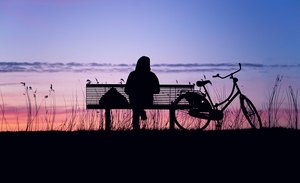 Es sitzt jemand alleine auf einer Bank am Meer und schaut in den rotgefärbten Abendhimmel, ein Fahrrad steht rechts neben der Bank