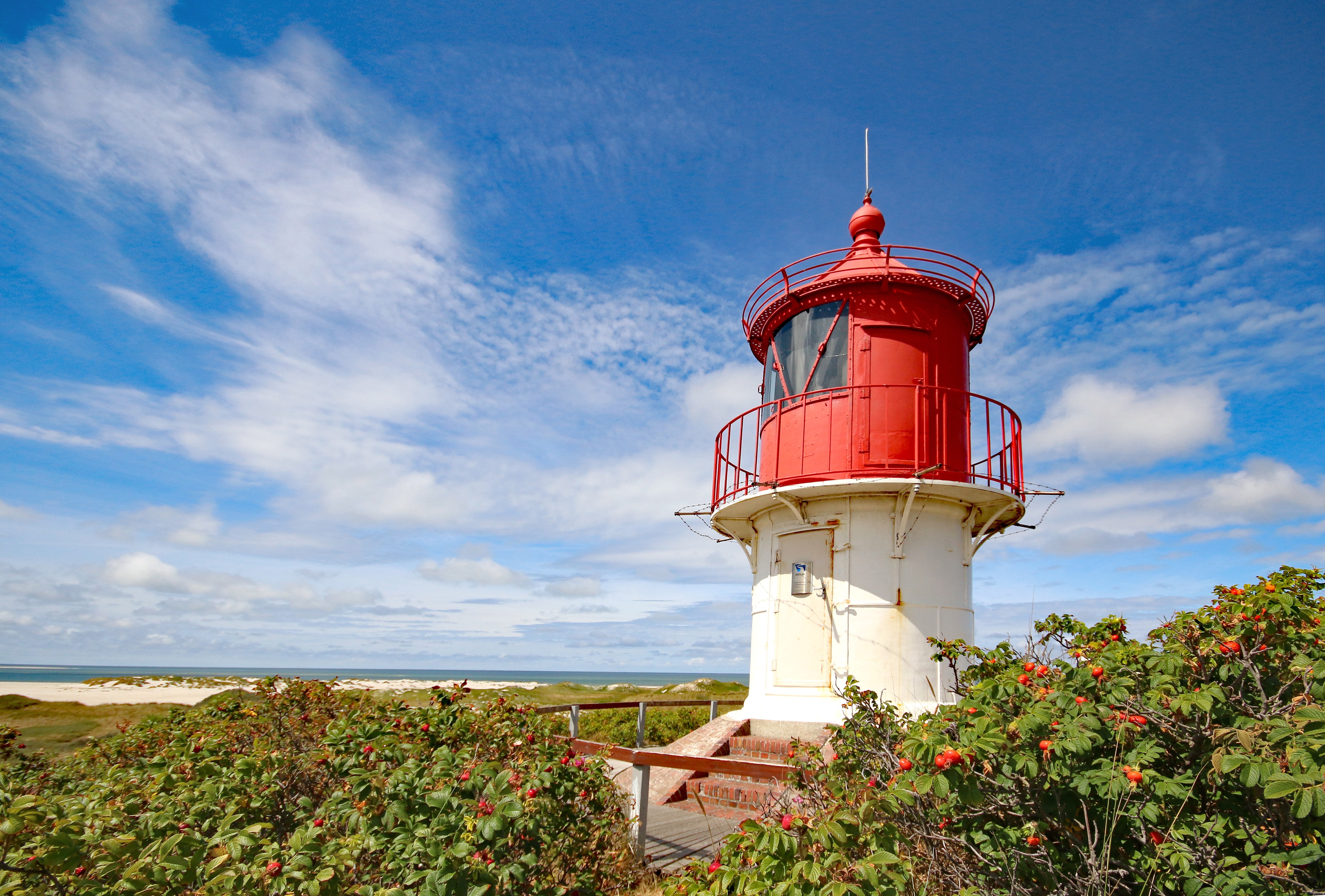 Zu sehen sind blauer Himmel über Amrum, ein kleiner Leuchtturm auf der rechten Bildseite sowie in einiger Entfernung Strand und Nordsee, davor Dünengras. 