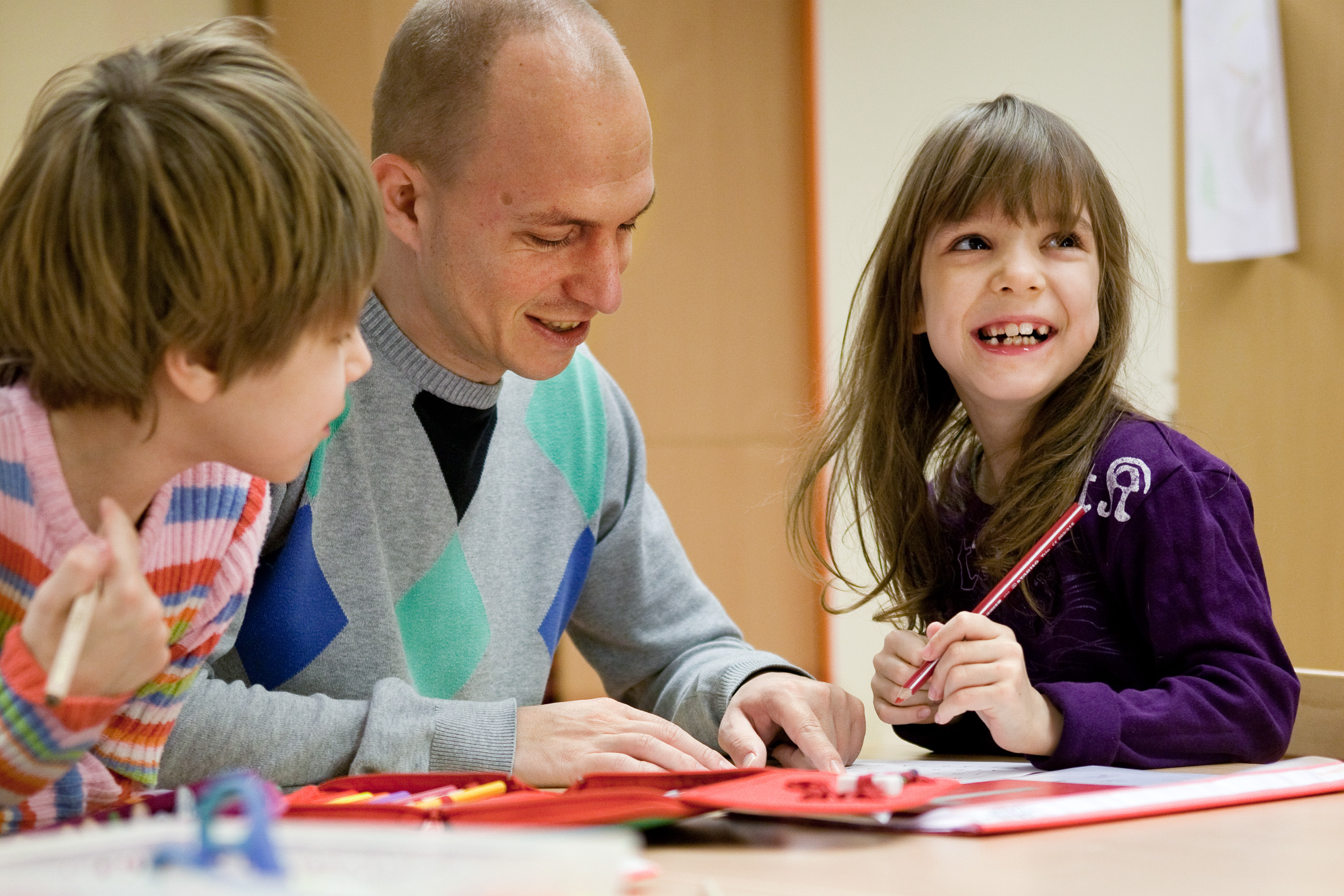 Ein BFDler sitzt mit zwei Kindern an einem Tisch und zeigt etwas in einer Unterlage, die Kinder sind interessiert nbzw. lachen.