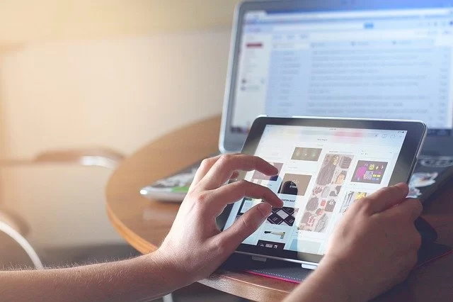 zwei Hände halten ein iPad, im Hintergrund ist eine  aufgeklapptes Laptop zu sehen
