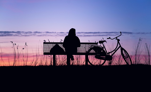 Es sitzt jemand alleine auf einer Bank am Meer und schaut in den rotgefärbten Abendhimmel, ein Fahrrad steht rechts neben der Bank